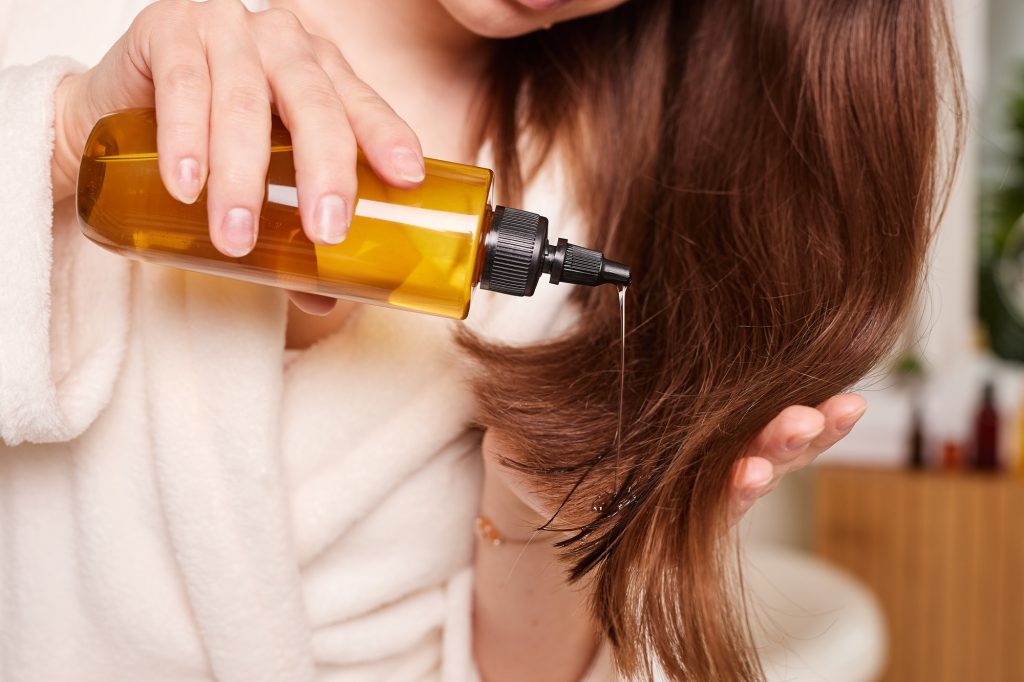 Femme appliquant de l'huile sur ses longs cheveux brillants sur fond de salle de bains, soins de santé capillaire