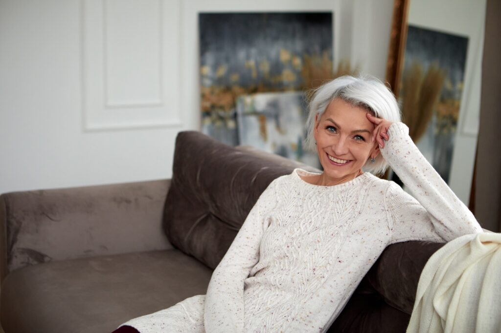 Charmante femme mature en pull tricoté et aux cheveux gris assise sur un canapé confortable et s'appuyant sur la main tout en regardant la caméra avec le sourire
