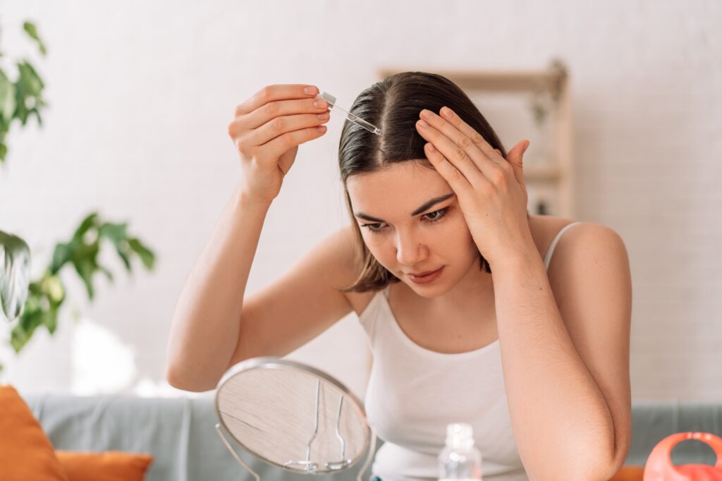 Une dame assise sur le canapé se regarde dans le miroir et verse de l'huile capillaire avec une pipette sur ses cheveux.
