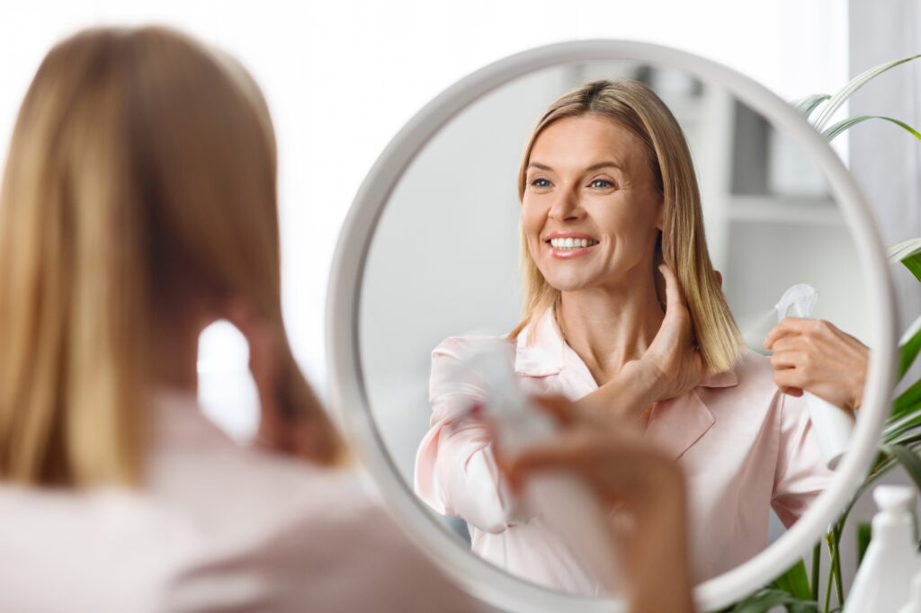 Belle femme d'âge moyen appliquant un fixatif hydratant sur les extrémités tout en étant assise près d'un miroir à la maison, une jolie dame d'âge mûr appréciant de nouveaux produits cosmétiques de coiffure et souriant à son reflet